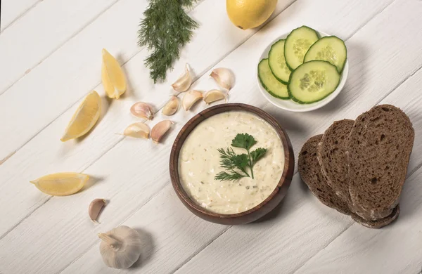 Top view of tzatziki sauce with greenery, cucumber and lemon on white wooden background — Stock Photo