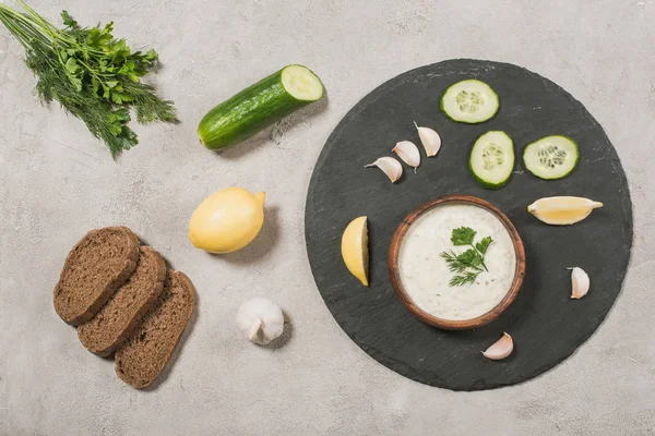 Вид сверху на соус хатцики с ингредиентами и хлебом на каменном фоне — стоковое фото