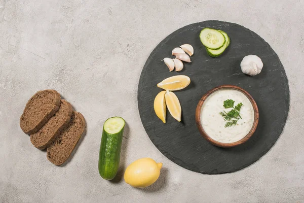 Vue du dessus de la délicieuse sauce tzatziki avec des ingrédients à bord sur fond de pierre — Photo de stock