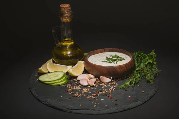 Bowl with tzatziki sauce with ingredients, spices and olive oil on dark board on black background — Stock Photo