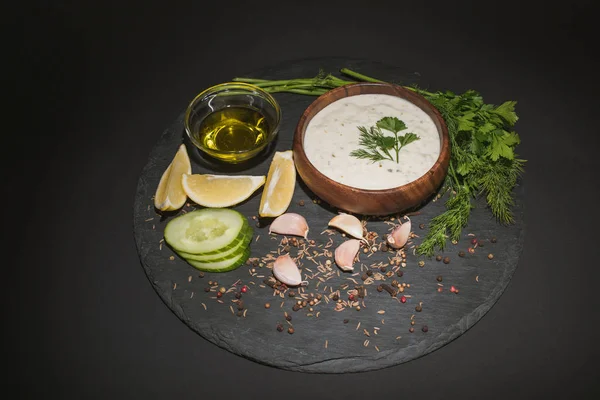 Leckere Tzatziki-Sauce mit Gewürzen, Olivenöl und Zutaten auf dunklem Brett auf schwarzem Hintergrund — Stockfoto