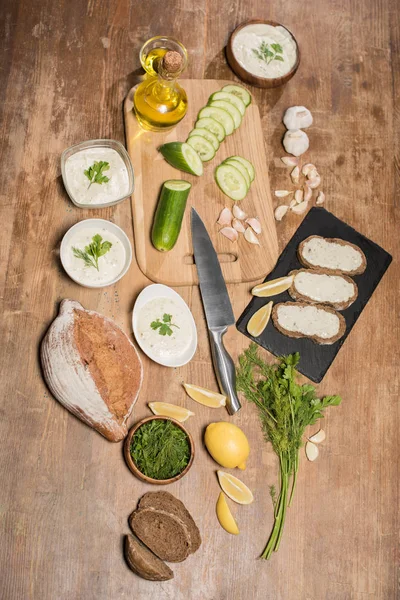 Вид сверху на соус хатцики с ингредиентами и хлебом на деревянном столе — стоковое фото