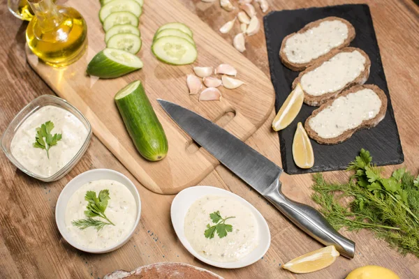 Top view of tzatziki sauce in bowls with bread and fresh ingredients on wooden table — Stock Photo