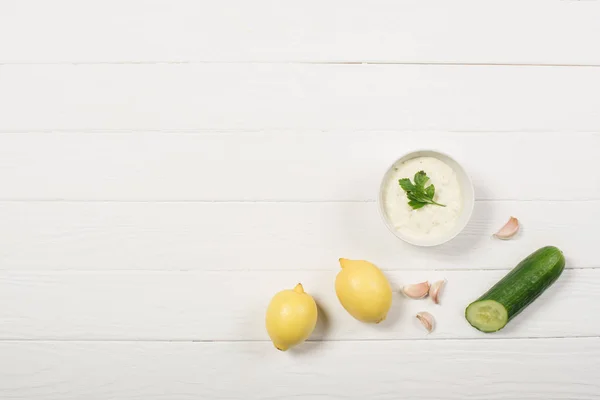 Vista superior de la salsa tzatziki con limones, pepino y ajo sobre fondo de madera blanca - foto de stock