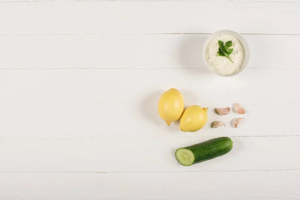 Draufsicht auf hausgemachte Tzatziki-Sauce mit Zitronen, Gurken und Knoblauch auf weißem Holzhintergrund — Stockfoto