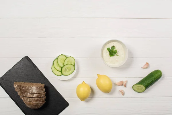 Vue du dessus de la sauce tzatziki avec du pain sur panneau noir sur fond blanc en bois — Photo de stock