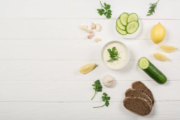 Вид зверху на соус Тцацікі інгредієнти, зелень і хліб на білому дерев'яному фоні — стокове фото