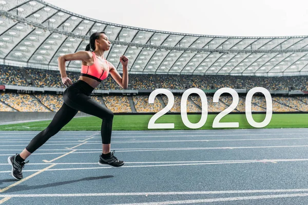 Jovem esportista afro-americana correndo no estádio perto de 2020 lettering — Fotografia de Stock