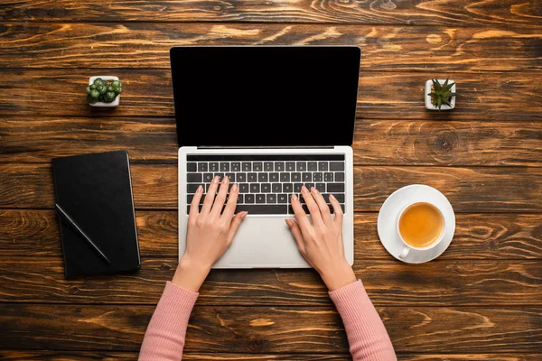 Cropped view of businesswoman using laptop near notebook, coffee cup and potted plants on wooden desk — Stock Photo