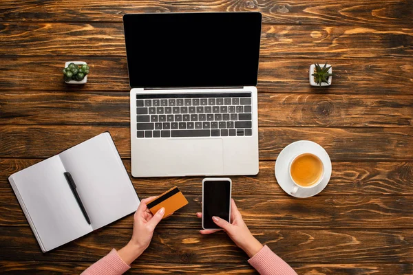 Vue recadrée de femme d'affaires tenant smartphone et carte de crédit près d'un ordinateur portable, ordinateur portable, tasse à café et plantes sur un bureau en bois — Photo de stock