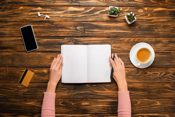 Partial view of businesswoman holding pen near blank notebook, coffee cup, smartphone, credit card, wireless earphones and plants on wooden desk — Stock Photo