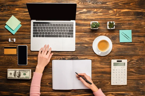 Vista recortada de la mujer de negocios escribiendo en un cuaderno cerca de la computadora portátil, teléfono inteligente, taza de café, tarjeta de crédito, dinero y calculadora en el escritorio de madera - foto de stock