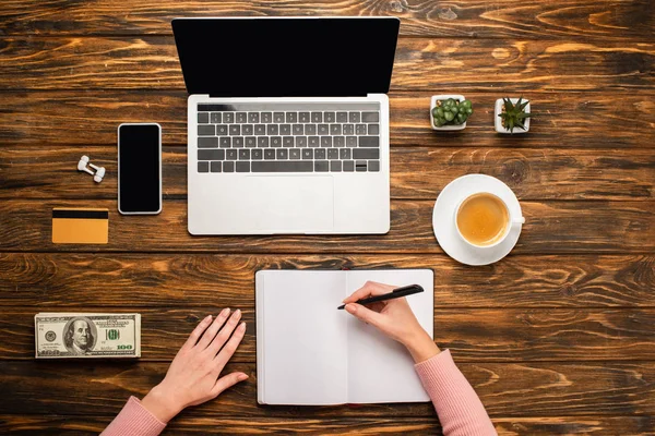 Vista recortada de la mujer de negocios escribiendo en un cuaderno cerca de la computadora portátil, teléfono inteligente, taza de café, tarjeta de crédito y billetes de dólar en el escritorio de madera - foto de stock