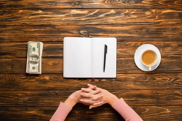 Обрізаний вид бізнес-леді, що розтягує руки під час підготовки до написання новорічної резолюції поблизу доларових банкнот і чашки кави на дерев'яному столі — стокове фото