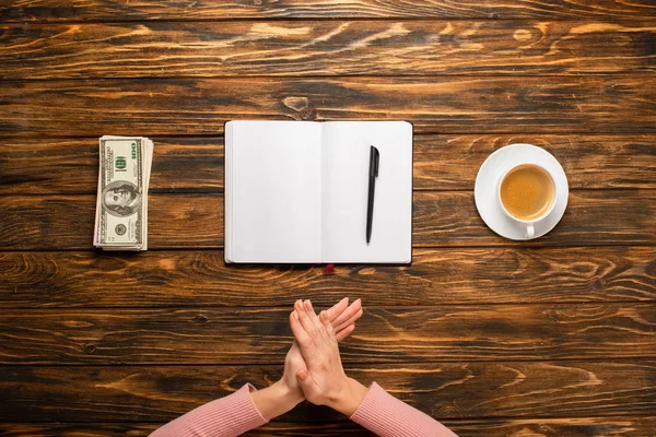 Abgeschnittene Ansicht einer Geschäftsfrau, die sich die Hände reibt, während sie sich darauf vorbereitet, Neujahrsvorsätze in der Nähe von Dollarnoten und Kaffeetasse auf Holztisch zu schreiben — Stockfoto