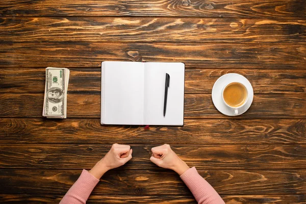 Vue recadrée de femme d'affaires tenant les poings tout en se préparant à écrire nouvelle résolution de l'année près de billets en dollars et tasse à café sur un bureau en bois — Photo de stock
