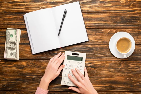 Vue recadrée de femme d'affaires à l'aide d'une calculatrice près d'un bloc-notes vierge, de billets en dollars et d'une tasse à café sur un bureau en bois — Photo de stock