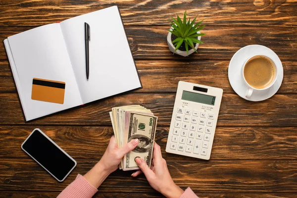 Частковий вид бізнес-леді, що рахує гроші поблизу калькулятора, смартфона, чашки кави, кредитної картки, ручки та блокнота на дерев'яному столі — стокове фото