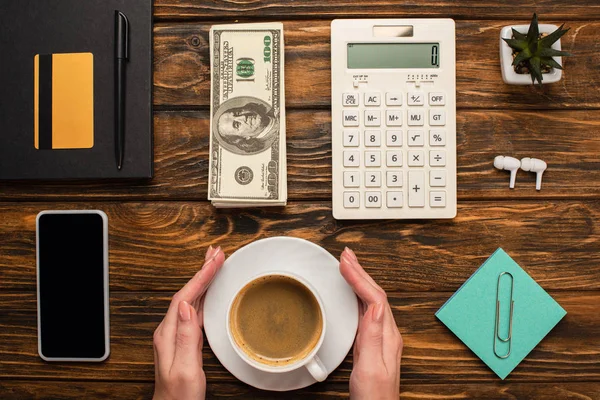 Обрезанный вид деловой женщины, держащей чашку кофе возле смартфона, долларовые купюры, калькулятор, беспроводные наушники и канцелярские принадлежности на деревянном столе — стоковое фото