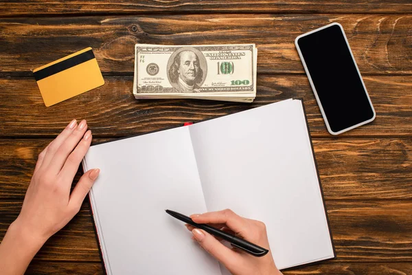 Vista recortada de empresária escrevendo em notebook em branco, smartphone, notas de dólar e cartão de crédito em mesa de madeira — Fotografia de Stock