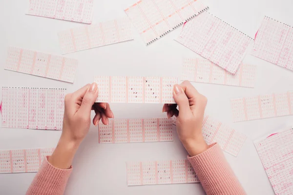 Vista ritagliata di donna in possesso di biglietto della lotteria con numeri segnati vicino a carte della lotteria sparse sul tavolo bianco — Foto stock