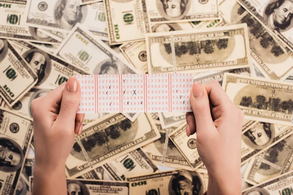 Vista recortada de la mujer sosteniendo billete de lotería con números marcados cerca de billetes de dólar dispersos - foto de stock