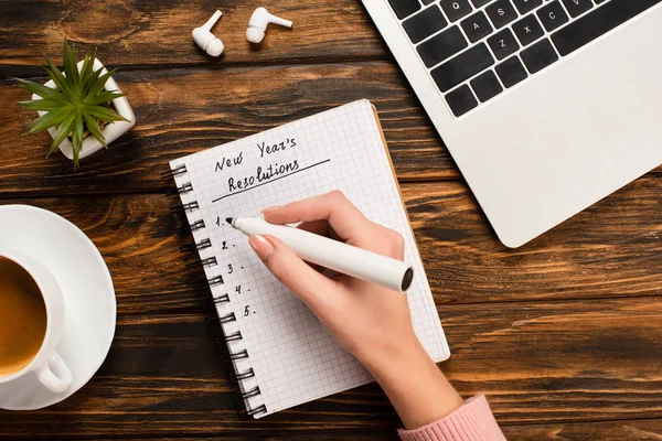 Vue partielle de femme d'affaires écrivant de nouvelles résolutions d'années près d'un ordinateur portable, écouteurs sans fil, plante en pot et tasse à café sur un bureau en bois — Photo de stock