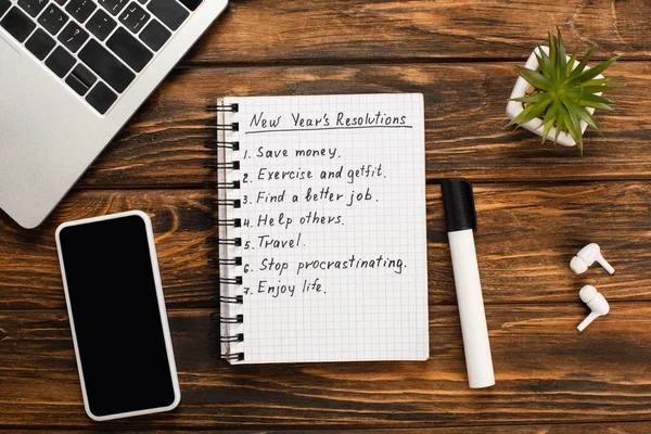 Notebook with list of new years resolutions, felt-tip pen, laptop, smartphone, potted plant and wireless earphones on wooden desk — Stock Photo