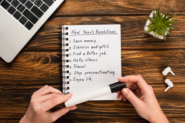 Cropped view of businesswoman holding felt-tip pen near notebook with list of new years resolutions near laptop, potted plant and wireless earphones on wooden desk — Stock Photo