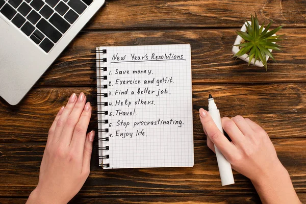 Cropped view of businesswoman holding felt-tip pen near notebook with list of new years resolutions near laptop and potted plant on wooden desk — Stock Photo