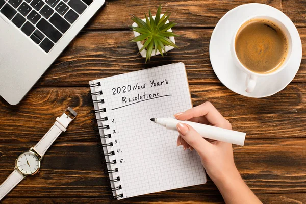 Cropped view of businesswoman holding felt-tip pen near notebook with 2020 new years resolution near coffee cup, laptop, wristwatch and potted plant on wooden desk — Stock Photo