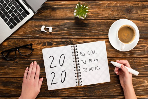 Cropped view of businesswoman holding felt-tip pen near notebook with 2020, goal, plan, action lettering near laptop, wireless earphones, coffee cup on wooden desk — Stock Photo