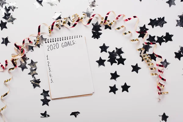 Top view of notebook with 2020 goals list with empty points near decorative, shiny stars and serpentine on white table — Stock Photo