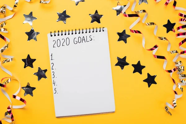 Notebook com metas 2020 com pontos numerados vazios perto de estrelas decorativas, brilhantes e serpentinas na superfície amarela — Fotografia de Stock