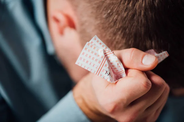 Selective focus of unhappy man sitting with bowed head while holding crumpled lottery ticket — Stock Photo