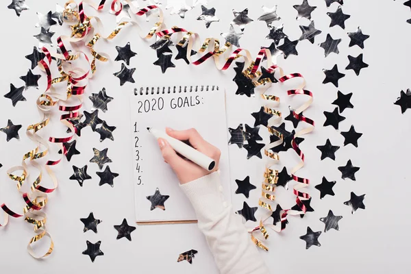 Vista recortada de la mujer de negocios escribiendo 2020 lista de objetivos en el cuaderno cerca de estrellas decorativas y serpentina en la mesa blanca - foto de stock