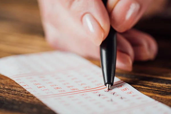 Close up view of hand of gambler marking numbers in lottery ticket with pen on wooden table — Stock Photo