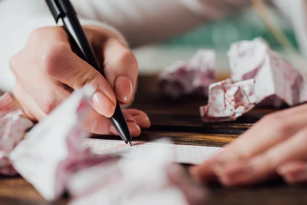 Ausgeschnittene Ansicht des Spielers, der Zahlen in Lottoschein neben zerknitterten Lottokarten auf Holztisch markiert — Stockfoto