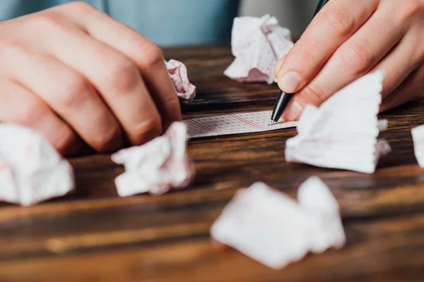 Vista parcial del jugador marcando números en billete de lotería cerca de cartas de lotería arrugadas en mesa de madera - foto de stock