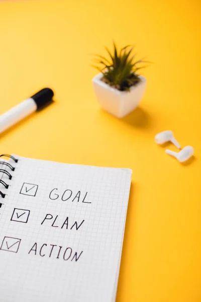 Notebook with goal, plan, action words near potted plant, wireless earphones and felt-tip pen on yellow desk — Stock Photo