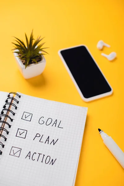 Notebook with goal, plan, action words near smartphone, potted plant, wireless earphones and felt-tip pen on yellow desk — Stock Photo