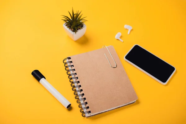 Notebook near smartphone with blank screen, wireless earphones, potted plant and felt-tip pen on yellow desk — Stock Photo