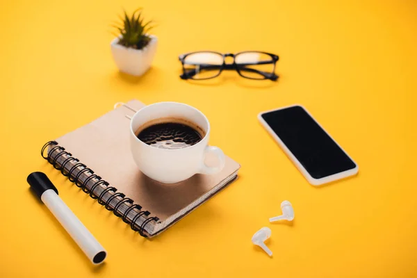 Coffee cup on notebook near smartphone, wireless earphones, glasses, potted plant and felt-tip pen on yellow desk — Stock Photo