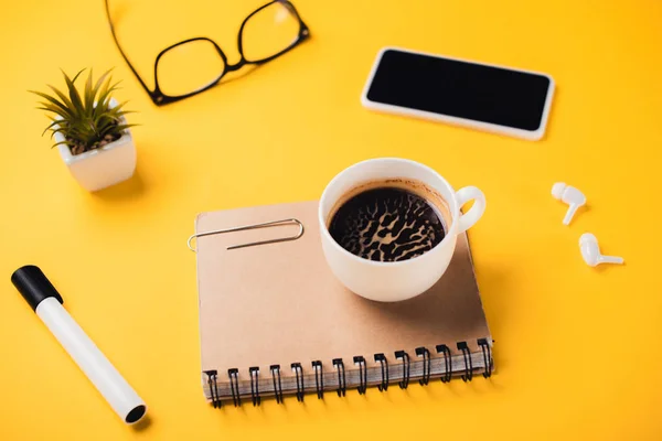 Coffee cup on notebook near smartphone, wireless earphones, glasses, potted plant and felt-tip pen on yellow desk — Stock Photo