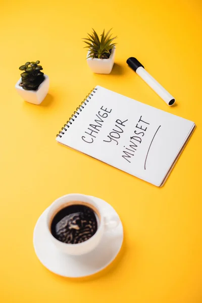 Cuaderno con cambio de mentalidad inscripción cerca de la taza de café, plantas en maceta y rotulador en el escritorio amarillo - foto de stock