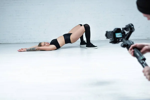 Selektiver Fokus des Kameramanns, der sexy blondes Mädchen im schwarzen Outfit auf dem Boden in der Nähe der weißen Ziegelwand dreht — Stockfoto