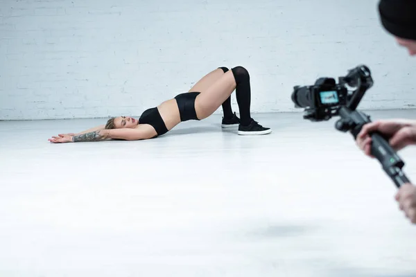 Enfoque selectivo de camarógrafo disparar sexy chica rubia en traje negro twerking en el suelo cerca de la pared de ladrillo blanco — Stock Photo