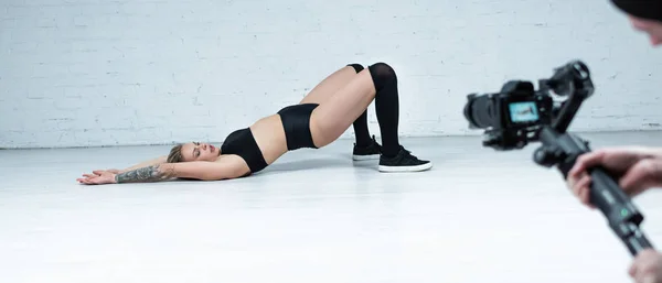 Selective focus of cameraman shooting sexy blonde girl in black outfit twerking on floor near white brick wall, panoramic shot — Stock Photo