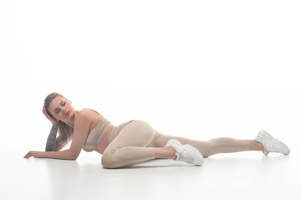 Sexy rubia chica en beige leggings twerking mientras se encuentra en el suelo aislado en blanco - foto de stock