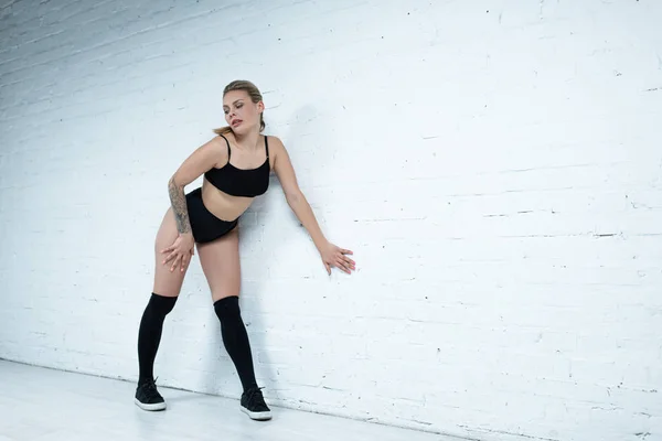 Tätowierte sexy blonde Mädchen im schwarzen Outfit twerking nahe Wand im Studio — Stockfoto